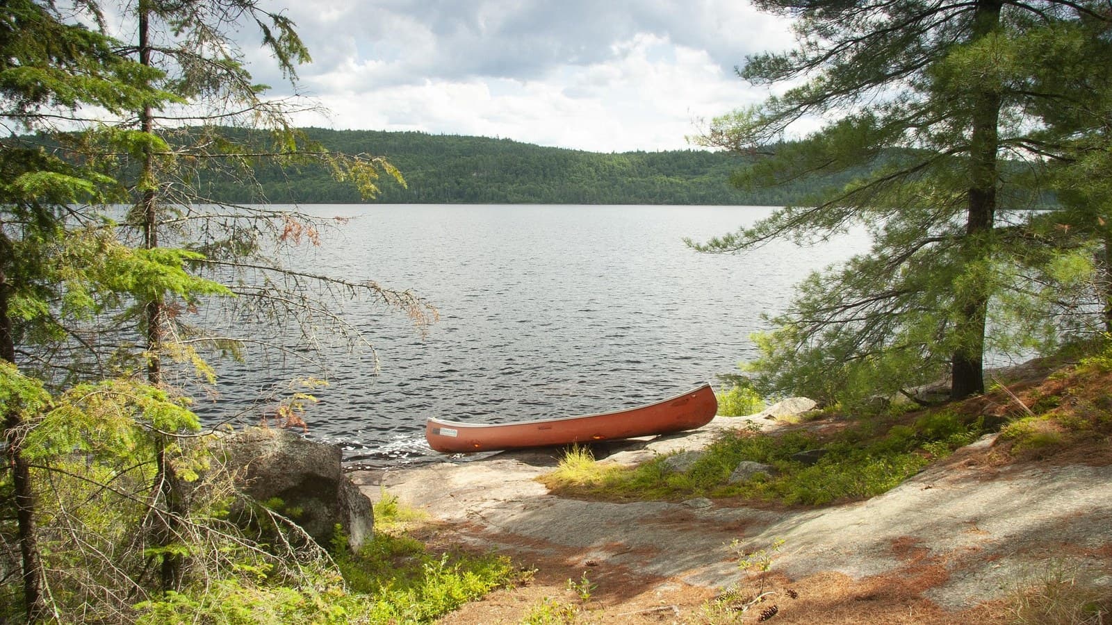 A red canoe pulled up on a granite shore beneath pines, with a wide lake and forested hills beyond.